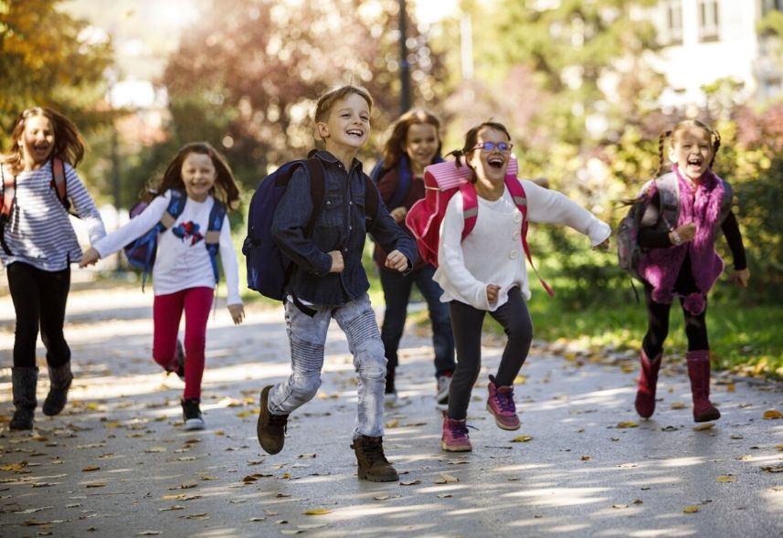 Campanha de matrícula: imagem de várias crianças correndo em uma rua com árvores ao fundo com mochilas nas costas.