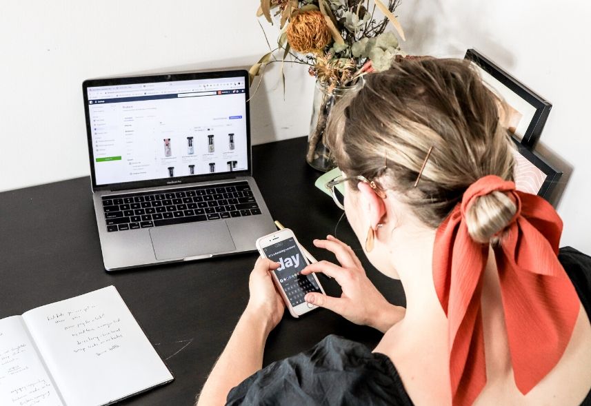 Como vender roupas pelo Instagram: imagem de uma mulher mexendo no celular com o notebook aberto em cima da mesa com imagens de roupas