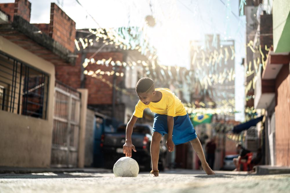 Dia das crianças nas redes sociais: imagem de um menino vestido de camiseta amarela e bermuda azul ajeitando uma bola no chão para brincar. A criança está na rua e algumas casas aparecem de fundo.
