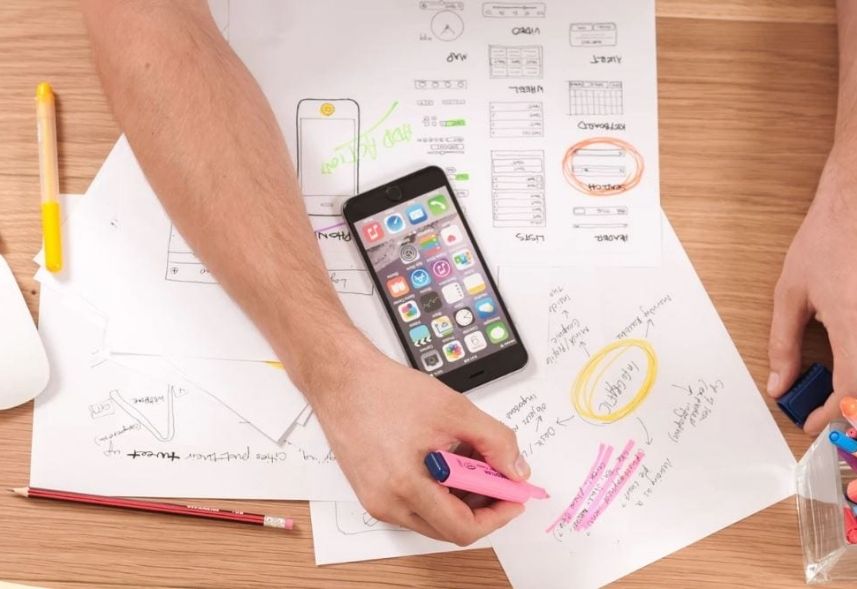 marketing digital para pequenas empresas: imagem de uma mão segurando uma caneta marca texto escrevendo em um papel com um celular ao lado sob uma mesa de madeira com várias anotações ao ao lado