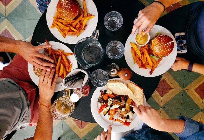Marketing para restaurantes: imagem de quatro pratos com comida e vários copos sob a mesa e as mãos das pessoas pegando os lanches e comidas